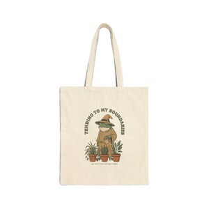 May include: Cream-colored tote bag featuring a whimsical illustration of a frog in a wizard's hat and robe, watering potted plants. Text reads "Tending to my boundaries" and "Do not touch the vibes". The bag has two handles.
