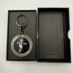 May include: A circular metal keyring featuring a colourful spaceship graphic against a black starry background, encased in a silver-toned frame with geometric detailing. The keyring is attached to a metal ring and rests inside a black box.