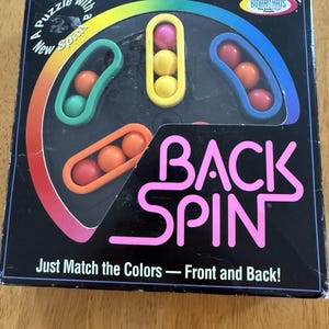 May include: A square game box for the Back Spin puzzle by Binary Arts. The black box features a rainbow arc, the text 'BACK SPIN', and 'Just Match the Colors — Front and Back!'. It displays a circular game board with colored balls in slots.
