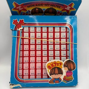 Könnte beinhalten: Ein blauer Pappkarton mit einem roten Kunststoffgitter und weißen Tasten, die mathematische Gleichungen zeigen. Die Schachtel trägt den Text Mon Aide-My Helper, zeigt Zeichentrickfiguren und hat ein gelbes Gummiband auf der Vorderseite.