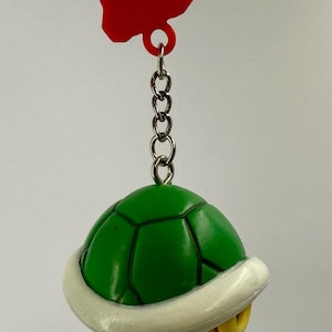 Puede incluir: Un llavero con forma de caparazón de tortuga verde con un borde blanco y una parte inferior amarilla. El caparazón está unido a una cadena de metal plateado y a una pieza de conexión de plástico rojo en la parte superior para colgar.