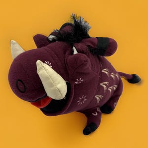 May include: A plush warthog toy in a deep maroon color with a soft fabric texture. It features large cream-colored tusks, a black tuft of hair on its head, and a red mouth. The body is decorated with small white embroidered star-like patterns.