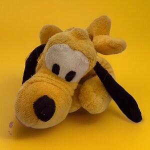 May include: A yellow plush dog toy with a white face, black ears, and a black nose, lying on a yellow background. The toy's design is soft and cuddly, with a classic cartoon aesthetic.