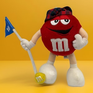 Puede incluir: Un peluche rojo de M&M's con un sombrero a cuadros y un palo de golf con una bandera azul con el número "18". El personaje está junto a una pelota de golf amarilla con el logotipo de M&M's.