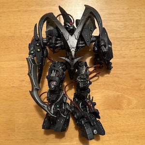 May include: A black and silver robotic action figure with intricate mechanical detailing, a pointed head crest, a red gem accent, and a large curved blade attached to its right arm. The figure features exposed tubing and complex armour plating.