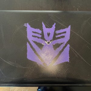 May include: Black laptop with a textured surface and a large purple emblem in the center. The emblem is a stylized, geometric design. The laptop shows signs of wear with visible scratches.