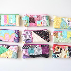May include: Nine colorful, quilted zipper pouches with patchwork designs. Each pouch features a unique combination of fabrics in various colors and patterns, with a small loop and zipper closure.