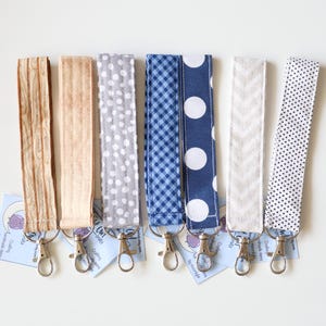 May include: A collection of seven fabric lanyards with various patterns, including wood grain, polka dots, and chevrons. Each lanyard has a silver-colored swivel clasp and a small tag. The lanyards are displayed on a white surface.