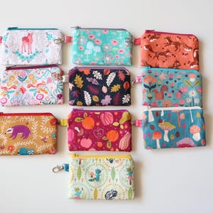May include: Nine small, zippered pouches with various colorful patterns. Designs include deer, flowers, foxes, leaves, fruit, and mushrooms. Each pouch has a small wrist strap and a silver-colored zipper pull.