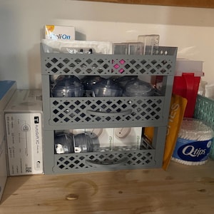 May include: A two-tiered grey plastic storage crate filled with various items. The top shelf holds several dark blue containers. The bottom shelf contains boxes with the text "dexcom G7". Other items include medical supplies and a container of Q-tips.