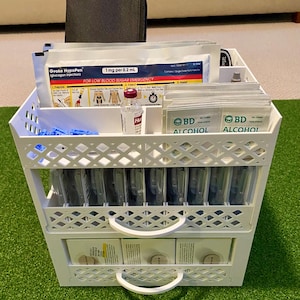 Op de afbeelding: Een witte, meerlaagse organizer gevuld met medische benodigdheden. De bovenste plank bevat Gvoke HypoPen, alcoholdoekjes en een flesje medicatie. De middelste plank bevat verschillende transparante, verpakte items. De onderste lade bevat extra medische benodigdheden. De tekst "FOR LOW BLOOD SUGAR EMERGENCY" is zichtbaar.