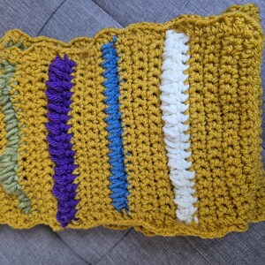May include: Mustard yellow crochet scarf with vertical stripes in white, blue, purple, and green. The scarf has a scalloped edge and a textured, handmade appearance. The crochet stitches are visible, adding to the handcrafted aesthetic.
