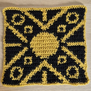 May include: A square crochet piece featuring a black background with a vibrant yellow geometric pattern. The design includes a central yellow circle, radiating diagonal lines, and small square and circular motifs in the corners and edges.