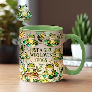 May include: A ceramic mug with a green handle and rim, featuring a stained-glass design of frogs on lily pads. The mug has the text "JUST A GIRL WHO LOVES FROGS" and the name "Sophia". A frog-shaped spoon rests on the rim.