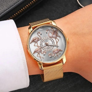 May include: Gold-toned wristwatch featuring a silver face with five flamingo designs. The watch has a gold mesh band and a gold bezel. The flamingo figures are embellished with sparkling accents. The watch is a stylish accessory.