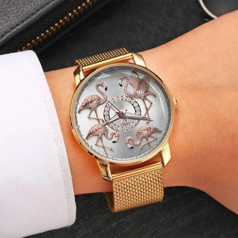 May include: Gold-toned wristwatch featuring a silver face with five flamingo designs. The watch has a gold mesh band and a gold bezel. The flamingo figures are embellished with sparkling accents. The watch is a stylish accessory.