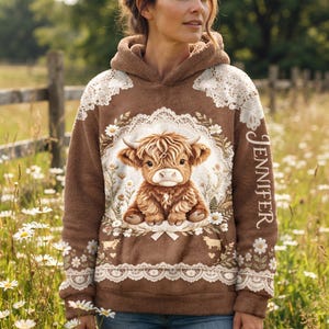 Puede incluir: Una sudadera con capucha de textura afelpada marrón que presenta un gráfico central de un ternero Highland rodeado de margaritas blancas y patrones de encaje. El nombre Jennifer está impreso verticalmente en blanco en el lado derecho.