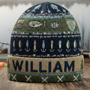 May include: A navy blue and olive green knit beanie featuring embroidered fishing motifs including fish, lures, and nets. The folded cuff is tan with the name WILLIAM in large navy blue letters, accented by small green pine tree patterns.