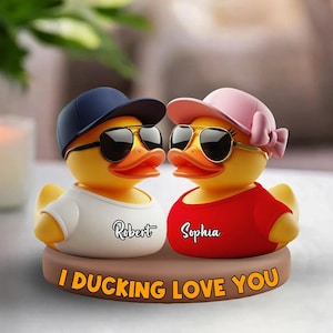 May include: Two yellow rubber duck figurines wearing sunglasses and hats. One duck wears a white shirt with "Robert" on it, the other a red shirt with "Sophia". They sit on a brown base with the text "I DUCKING LOVE YOU".