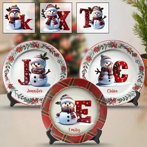 Può includere: Piatti decorativi e ornamenti con lettere a tema natalizio con pupazzi di neve. I piatti hanno uno sfondo bianco con un bordo floreale rosso. Ogni piatto mostra un pupazzo di neve accanto a una lettera rossa e un nome. Le lettere sono K, X, T, J, C ed E.