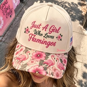 Puede incluir: Una gorra de béisbol beige con el texto bordado Just A Girl Who Loves Flamingos en rosa, acentuado por pequeñas flores rosas. La visera muestra un patrón de flamencos rosas e hibiscos. Un pequeño parche de la bandera estadounidense está en el lateral.
