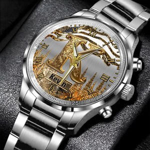 May include: Silver wristwatch with a detailed face featuring a gold-colored letter "K" and the name "Kevin". The watch has a silver band and a decorative background with gold and white accents. Roman numerals mark the hours.