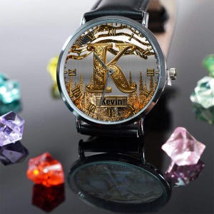 May include: A personalized wristwatch with a black leather band. The watch face features a gold-colored letter "K" with an ornate design, Roman numerals, and the name "Kevin" displayed. The watch is set against a backdrop of a forest scene.
