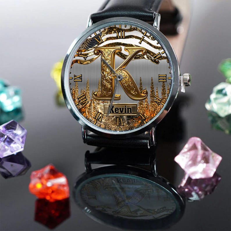 May include: A personalized wristwatch with a black leather band. The watch face features a gold-colored letter "K" with an ornate design, Roman numerals, and the name "Kevin" displayed. The watch is set against a backdrop of a forest scene.