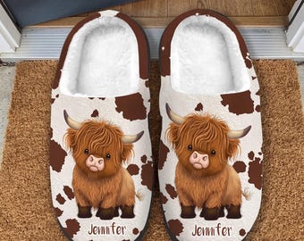 Puede incluir: Un par de pantuflas con un patrón de manchas de vaca blancas y marrones. Cada pantufla muestra un gráfico de una vaca Highland marrón y peluda con cuernos y el nombre Jennifer impreso en letras cursivas marrones debajo.
