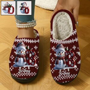 May include: Burgundy and white patterned slippers featuring a snowman design. The snowman wears a hat and scarf, with a red letter "B" and the name "Britney" printed on the slipper. The interior is lined with soft, cream-colored material.