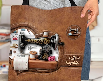 Peut inclure: Sac fourre-tout marron avec un motif de machine à coudre vintage. Le sac a une texture semblable à du cuir avec une illustration détaillée d'une machine à coudre noire et dorée, des bobines de fil et d'autres accessoires de couture. Le nom "Sophia" est également visible.