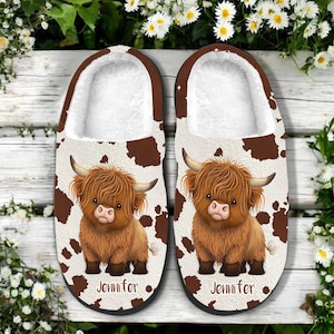 May include: A pair of cozy, open-toe slippers featuring a brown and white cowhide pattern. Each slipper displays a cartoon illustration of a Highland cow with the name "Jennifer" printed below. The interior is lined with soft, white faux fur.
