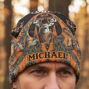 May include: A black, orange, and brown beanie featuring a deer graphic and the words "DEER HUNTING" and "MICHAEL". The beanie has a camouflage pattern and is worn by a person.