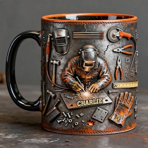 Può includere: Una tazza in ceramica con manico nero e bordo arancione, raffigurante un saldatore con casco e guanti che lavora su una piastra metallica con la scritta CHARLIE. Il design include vari strumenti come pinze, una smerigliatrice e un martello.