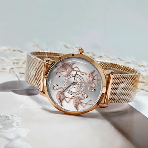 May include: A rose gold wristwatch with a mesh band. The watch face is silver with rose gold flamingo designs and a diamond-studded center. The watch has a classic analog display.