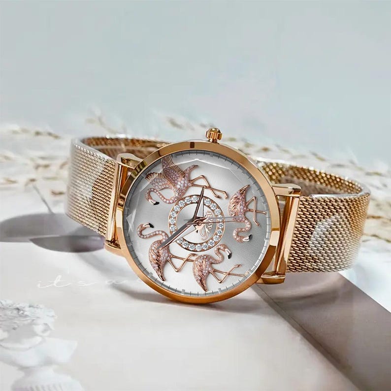 May include: A rose gold wristwatch with a mesh band. The watch face is silver with rose gold flamingo designs and a diamond-studded center. The watch has a classic analog display.