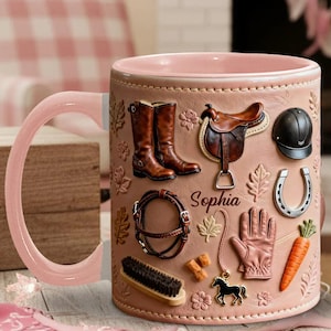 May include: A pink ceramic mug with a horse theme. The mug features embossed images of riding boots, a saddle, a helmet, a horseshoe, a glove, a carrot, and a horse charm. The name "Sophia" is written on the mug.
