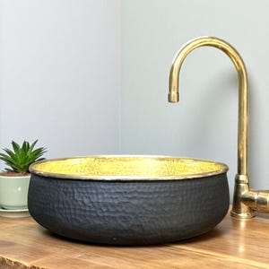 May include: A black hammered metal vessel sink with a gold interior and rim. A gold faucet is positioned to the right. A small potted succulent plant sits to the left on a wooden surface.