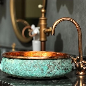 May include: A copper bathroom sink with a verdigris finish and a copper rim. The sink is paired with a copper faucet and fixtures. The sink is set against a dark gray wall with a gold-framed mirror.