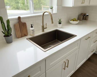 Puede incluir: Una cocina moderna con encimera y armarios blancos, y un fregadero marrón oscuro. Un grifo y herrajes dorados complementan el diseño. Los elementos decorativos incluyen una tabla de cortar de madera, plantas en macetas y un dispensador de jabón.