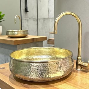 May include: A hammered gold-colored sink basin with a matching faucet. The basin has a textured surface and sits on a wooden countertop. A smaller gold basin and faucet are visible in the background, along with a small potted plant.