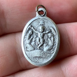 K&ouml;nnte beinhalten: Silberner ovaler Anh&auml;nger mit einem Reliefbild des Jesuskindes, umgeben von zwei Engeln. Die Inschrift lautet "O Infant Jesus Protect Us".