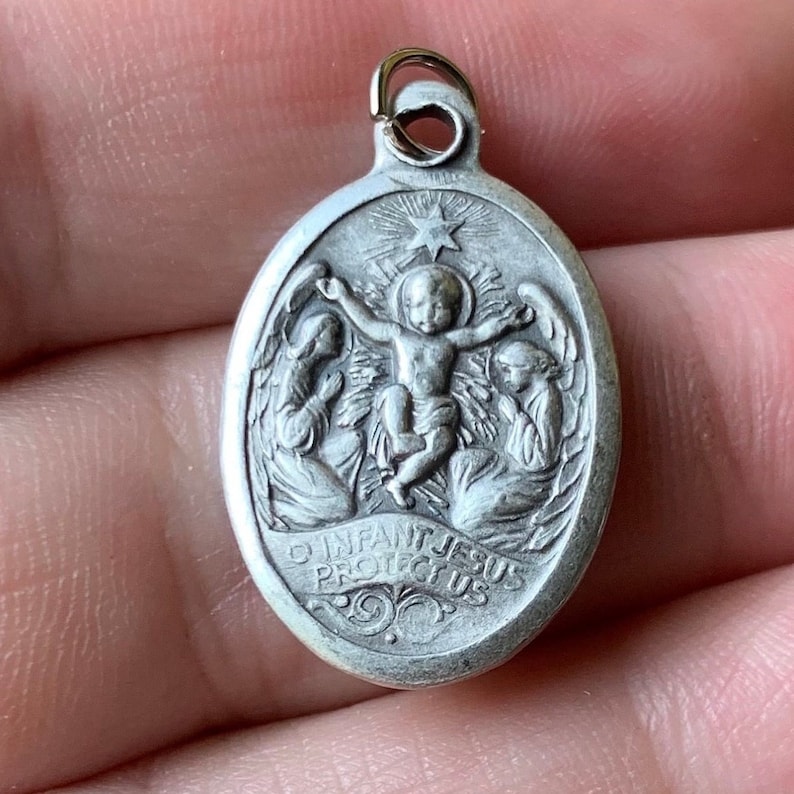K&ouml;nnte beinhalten: Silberner ovaler Anh&auml;nger mit einem Reliefbild des Jesuskindes, umgeben von zwei Engeln. Die Inschrift lautet "O Infant Jesus Protect Us".