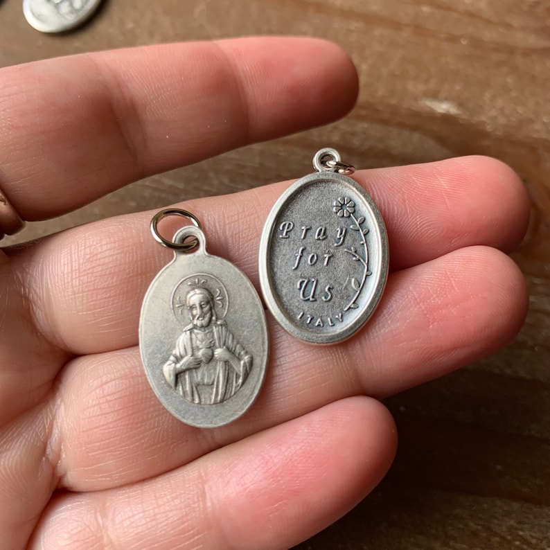 K&ouml;nnte beinhalten: Zwei silberne ovale Anh&auml;nger. Ein Anh&auml;nger zeigt die Darstellung eines Mannes mit einem Herzen in der Hand. Der andere Anh&auml;nger tr&auml;gt die Worte "Pray for Us" und ein kleines Blumenmuster. Beide Anh&auml;nger haben "Italy" auf der R&uuml;ckseite.