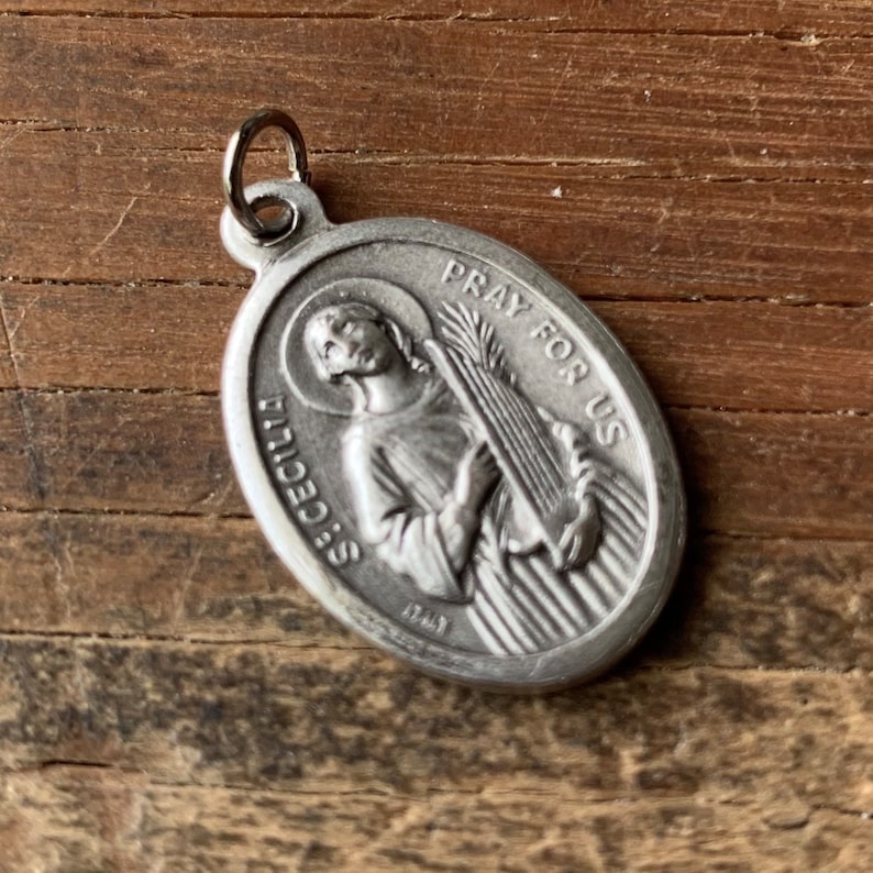 K&ouml;nnte beinhalten: Silberner ovaler Anh&auml;nger mit einer Darstellung der Heiligen C&auml;cilia, die ein Musikinstrument h&auml;lt. Der Anh&auml;nger tr&auml;gt die Inschriften "Pray for Us" und "St. Cecilia".