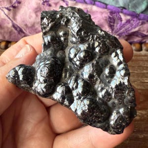 May include: A dark, metallic-looking mineral specimen with a bumpy, textured surface. The color is a deep, reflective black, with areas that catch the light, creating highlights. The overall shape is irregular and organic.