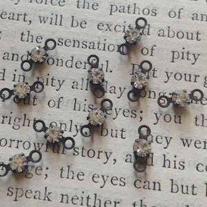 May include: Close-up of several small, dark metal connectors with a clear, faceted gem in the center. Each connector has two small loops on either side of the gem. The connectors are scattered on a vintage book page.