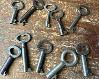 May include: A collection of nine antique-style metal keys with varying bow shapes and shaft lengths. Each key features a distinct bit pattern at the end of the shaft and a circular or oval head, all finished in a weathered, dark gray metallic tone.