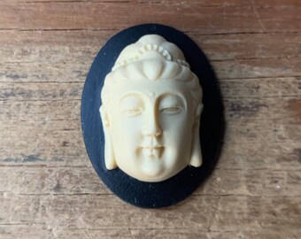 May include: A white ceramic cameo of a Buddha face, set in a black oval frame.