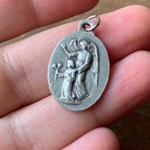 K&ouml;nnte beinhalten: Silberner ovaler Anh&auml;nger mit einem detaillierten Relief eines Engels mit Fl&uuml;geln, der die Hand eines kleinen Kindes h&auml;lt. Der Engel tr&auml;gt eine flie&szlig;ende Robe und das Kind h&auml;lt einen Blumenstrau&szlig;.